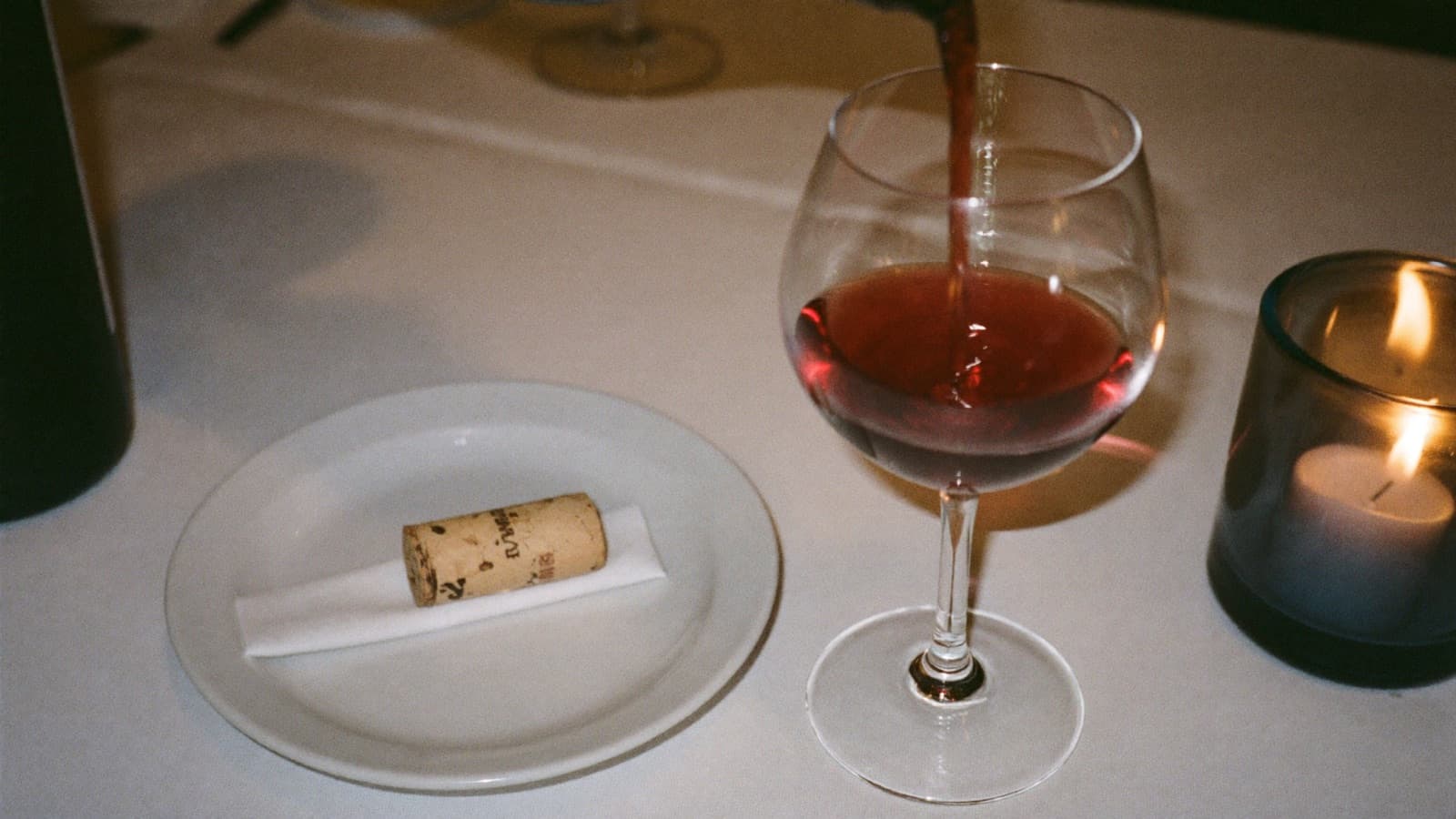 A server presenting a wine cork to a seated guest beside a small taste pour