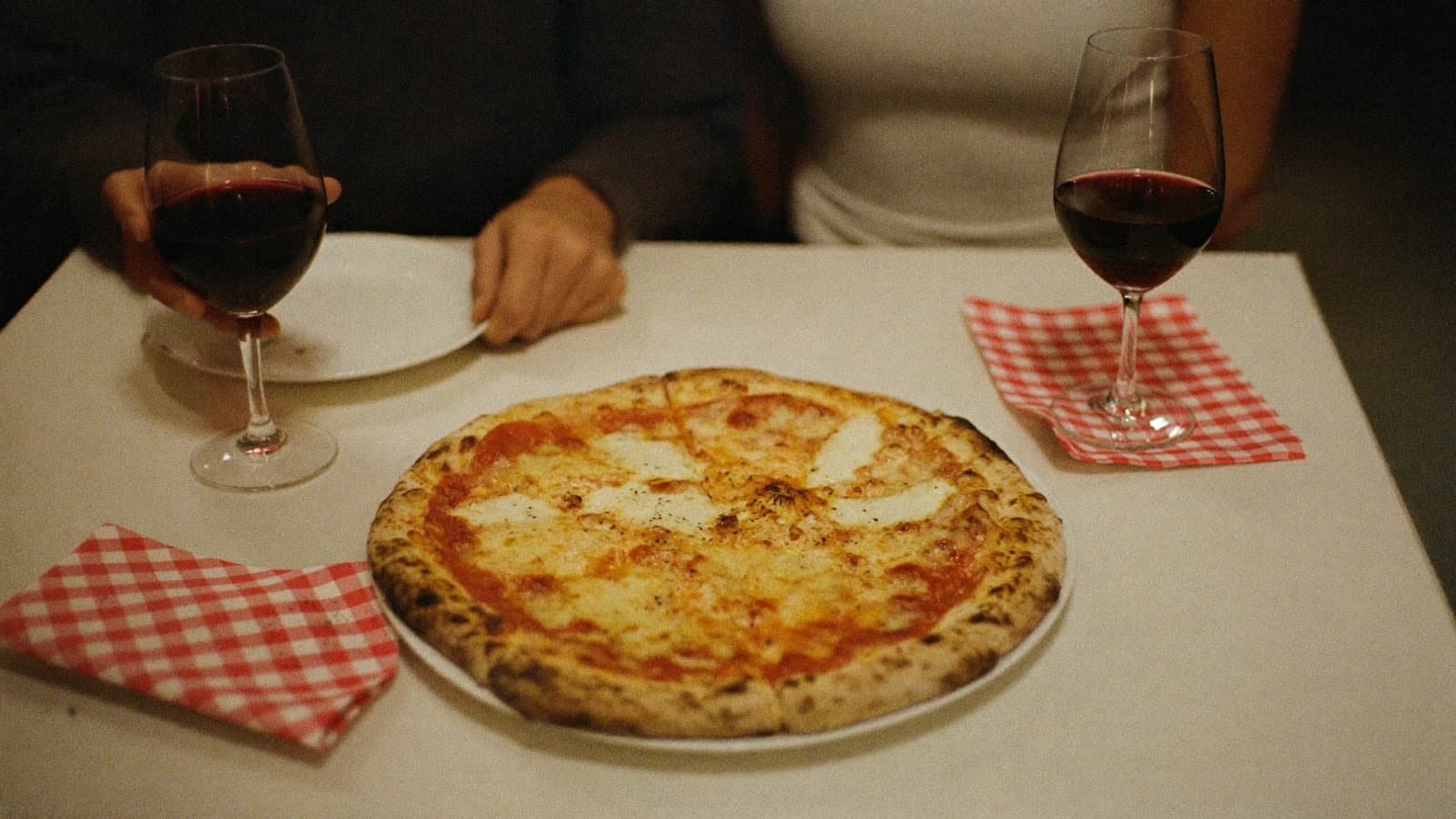 A margherita pizza with two tumblers of red wine on a casual checked-napkin table
