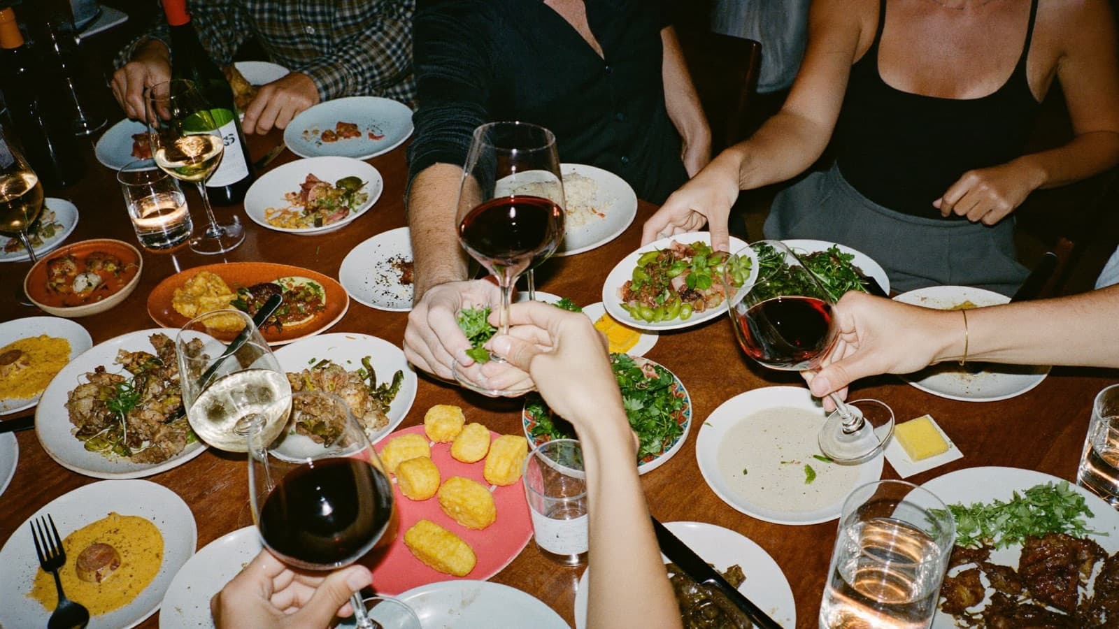 A lively shared dinner table of small plates and several wine glasses, hands passing dishes