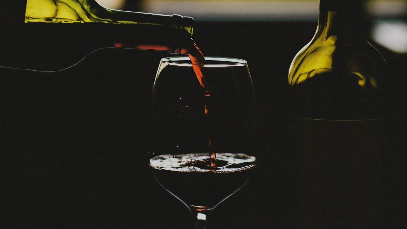 A single glass pour of red wine beside a full bottle on a warm bar counter