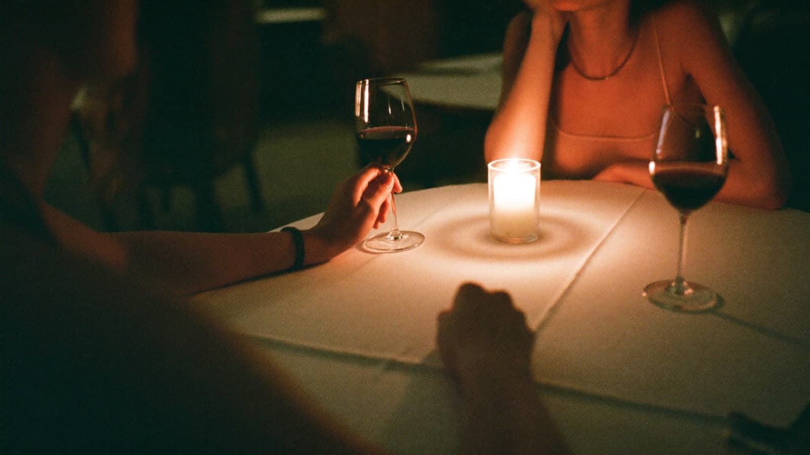 A candlelit table for two with glasses of red wine and two hands almost touching