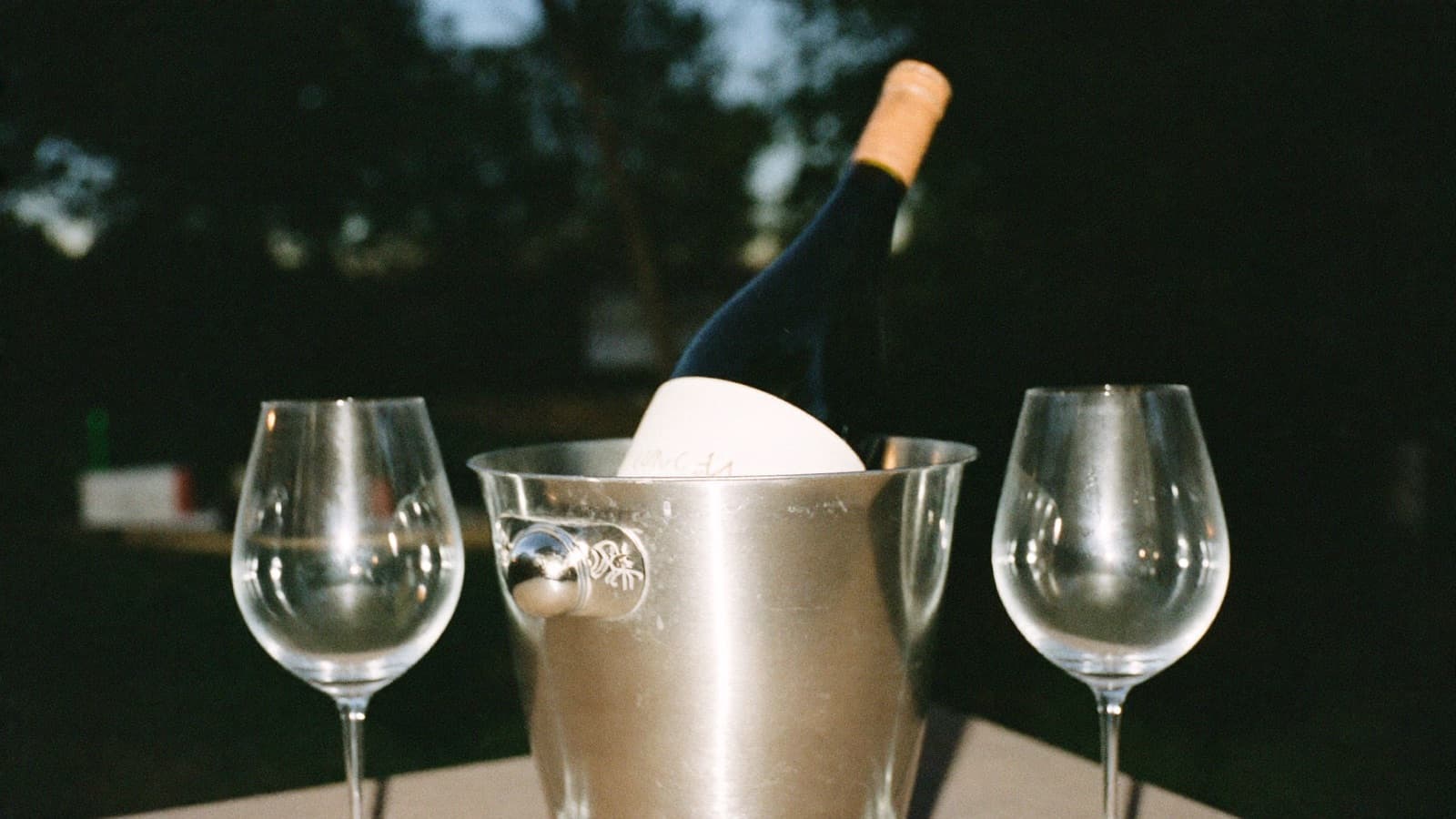 A bottle of white wine chilling in an ice bucket beside two empty glasses on a patio
